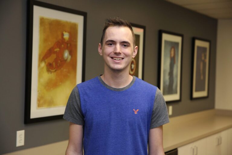 A young man wearing a blue and gray t-shirt stands indoors, smiling in front of a wall with four framed artworks.