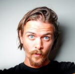 A man with light skin, blue eyes, and a mustache with a goatee looks surprised, facing the camera against a plain light gray background.