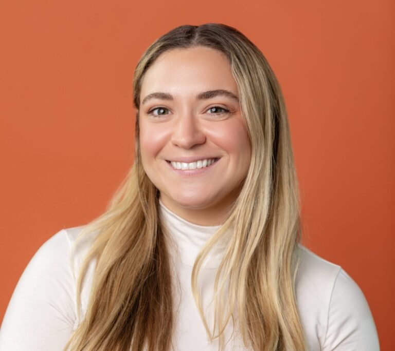 A woman with long blonde hair, wearing a light-colored turtleneck, smiles in front of an orange background.