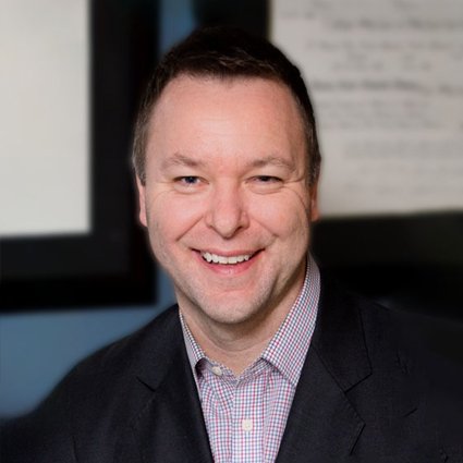 A man in a dark blazer and checked shirt smiles at the camera, with blurred framed documents or artwork in the background.