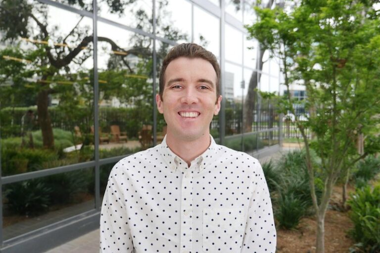 A man wearing a white shirt with black polka dots stands outside in front of a building with large glass windows and greenery.