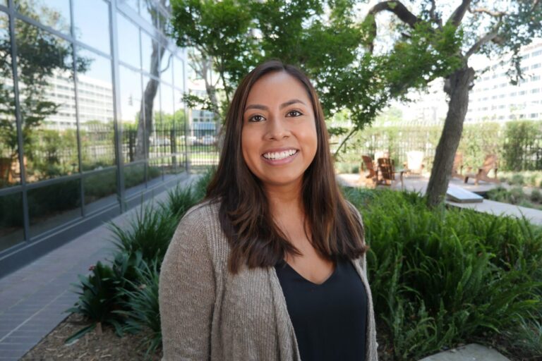 A woman with long brown hair smiles at the camera while standing outdoors next to a building and greenery.