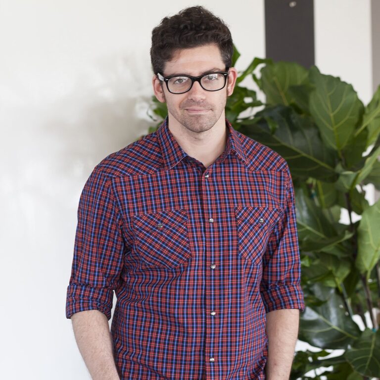 A man wearing glasses and a red plaid shirt stands in front of a green leafy plant, looking at the camera with a neutral expression.