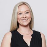 A woman with long blonde hair, wearing a sleeveless black top and a gold necklace, smiles at the camera against a plain light background.