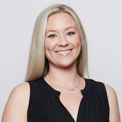 A woman with long blonde hair, wearing a sleeveless black top and a gold necklace, smiles at the camera against a plain light background.