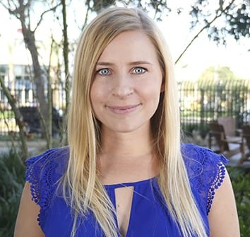 A woman with straight blonde hair wearing a blue blouse stands outdoors in front of trees and a fence, looking at the camera and smiling slightly.