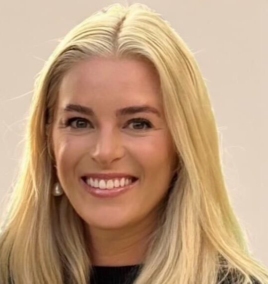 A woman with long blonde hair, wearing a black top, smiles at the camera against a neutral background.