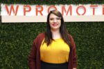 A woman with long dark hair and glasses, wearing a yellow top and maroon cardigan, stands in front of a green leafy wall with a partial "WPROMOTE" sign.