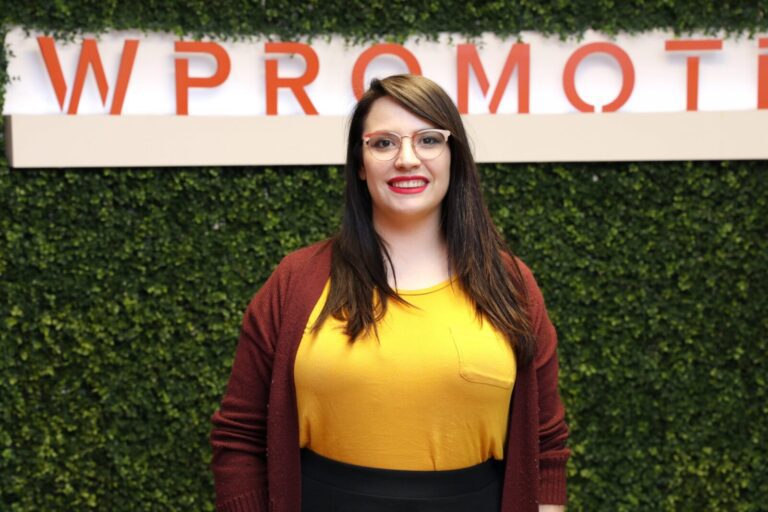 A woman with long dark hair and glasses, wearing a yellow top and maroon cardigan, stands in front of a green leafy wall with a partial "WPROMOTE" sign.