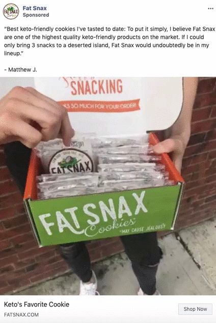 Person holding a box of individually wrapped Fat Snax cookies with a sign behind that reads "SNACKING"; box displays the brand name and slogan.