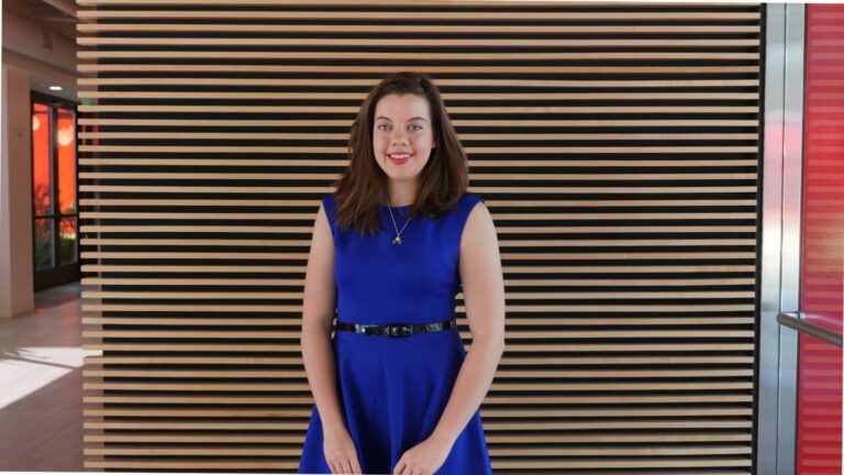 A woman in a blue dress stands in front of a modern wooden slat wall, smiling at the camera in a brightly lit indoor space.