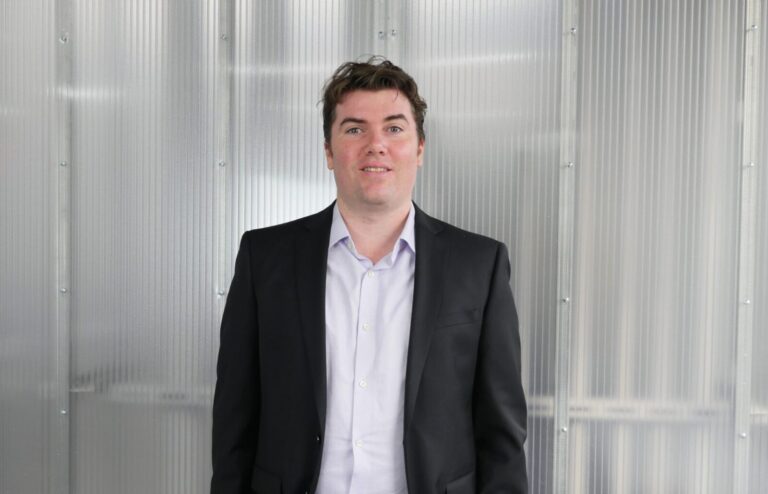 A man in a dark suit jacket and white shirt stands in front of a metallic, corrugated wall, facing the camera with a neutral expression.