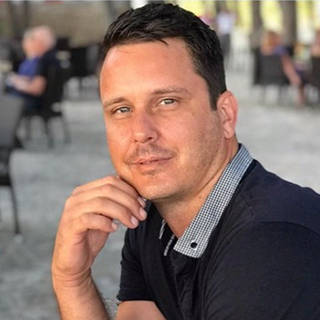 A man with short dark hair and a navy collared shirt sits outdoors, resting his chin on his hand, with blurred people and tables in the background.