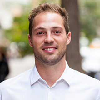 A man with short light brown hair and a trimmed beard is smiling, wearing a white collared shirt, standing outdoors with trees in the background.