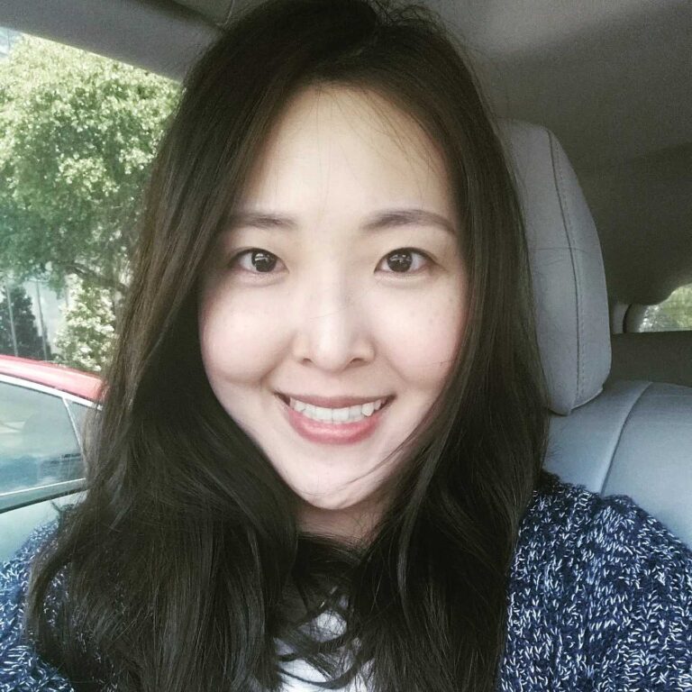 A woman with long dark hair sits in a car, smiling at the camera. Trees and a red car are visible through the window in the background.