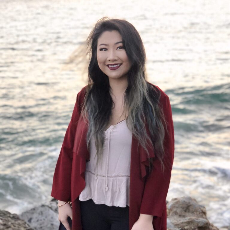 A woman with long dark hair and a red jacket stands on rocks by the ocean, smiling at the camera with waves in the background.
