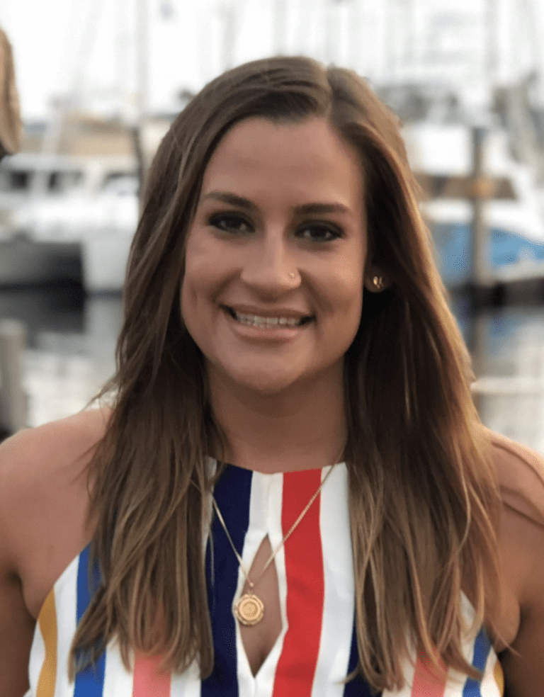 A woman with long brown hair and a striped sleeveless top smiles at the camera, with boats and water in the background.