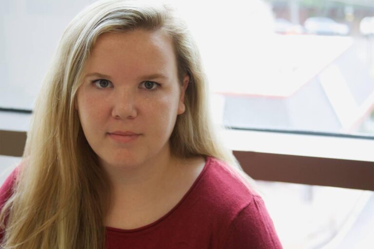 A woman with long blonde hair and a red shirt sits indoors near a window, looking directly at the camera.