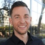 A man with short brown hair and a trimmed beard smiles outdoors in front of a glass building reflecting trees and city elements.