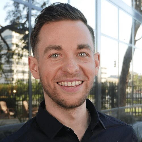 A man with short brown hair and a trimmed beard smiles outdoors in front of a glass building reflecting trees and city elements.