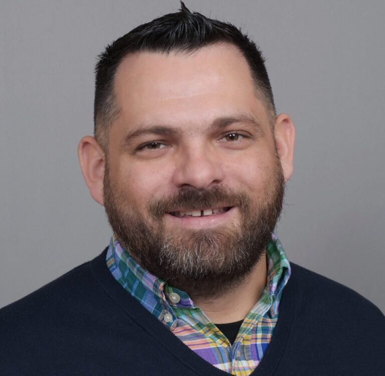 Man with short dark hair and beard, wearing a multicolored plaid shirt under a dark sweater, smiles at the camera against a plain gray background.
