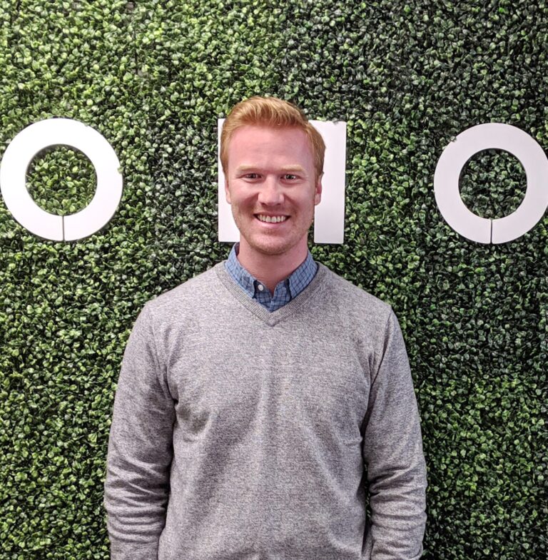 A man with short blond hair smiles while standing in front of a green artificial plant wall with large white letters.