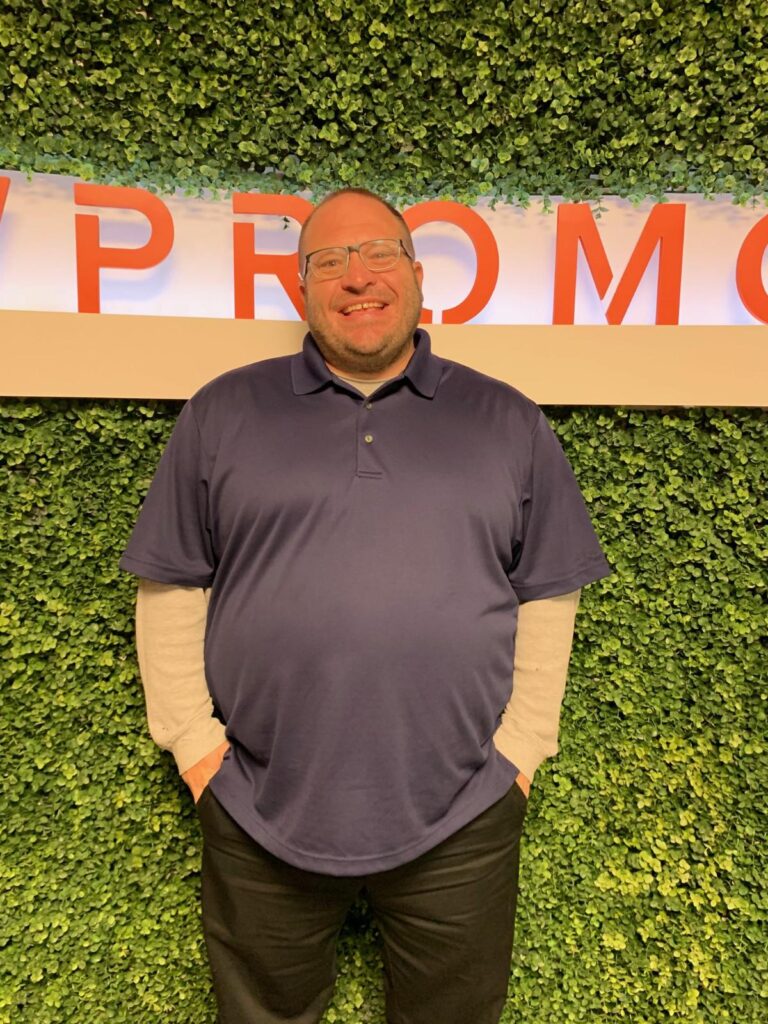 A man in a blue polo shirt and glasses stands in front of a green leafy wall with part of a sign reading "PROMO" visible above him.