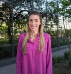 A woman with long straight hair wearing a magenta button-up shirt stands outside in front of a glass building and greenery, smiling at the camera.