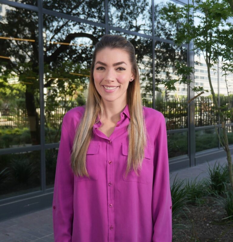 A woman with long straight hair wearing a magenta button-up shirt stands outside in front of a glass building and greenery, smiling at the camera.