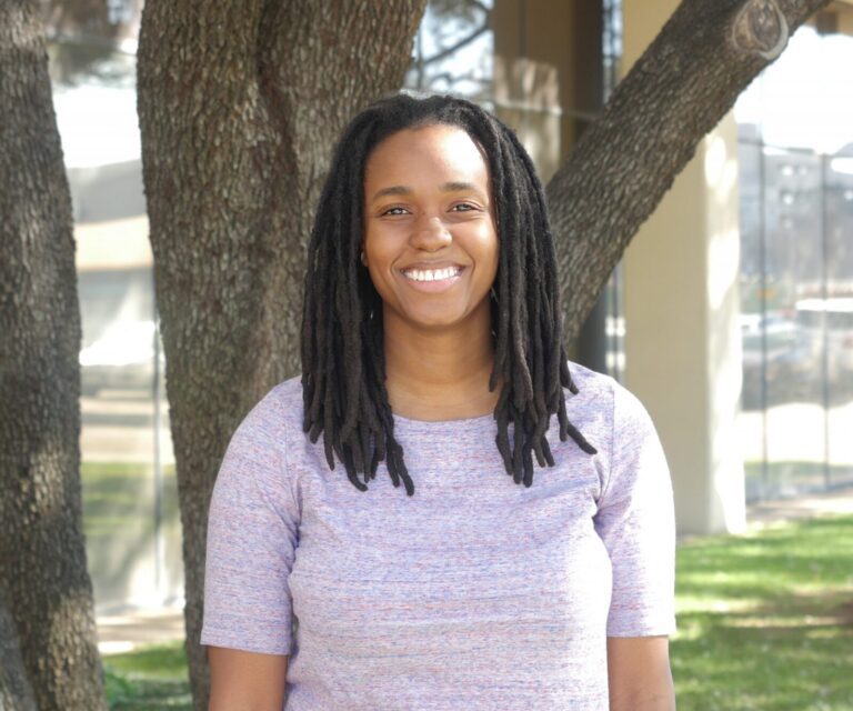 A person with long, dark dreadlocks and a light purple shirt stands outdoors in front of trees, smiling at the camera.