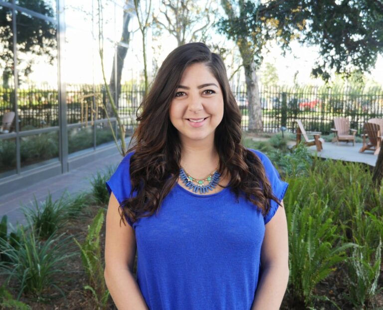 Woman with long brown hair wearing a blue shirt and necklace, standing outdoors in a garden area with chairs and trees in the background.