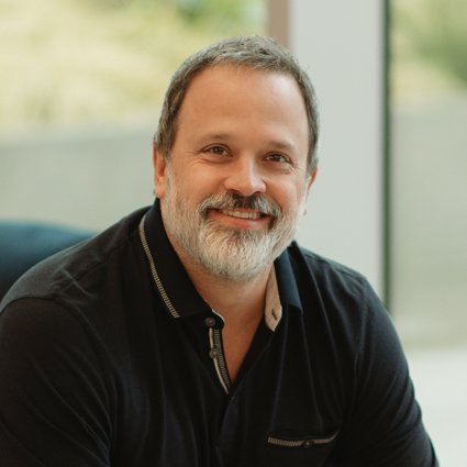 A middle-aged man with short dark hair and a gray beard, wearing a dark collared shirt, smiles while seated indoors near a large window.