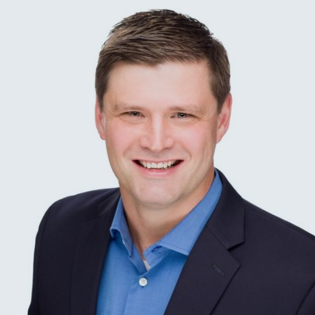 Man wearing a navy blazer and blue shirt, smiling, with short brown hair, posed against a plain light background.