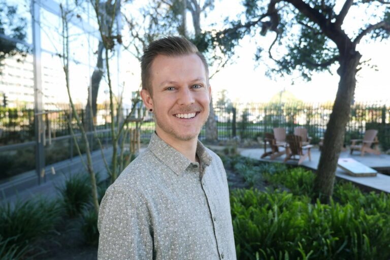 A man with short hair and a light patterned shirt smiles outdoors in a garden area with trees, greenery, and wooden chairs in the background.