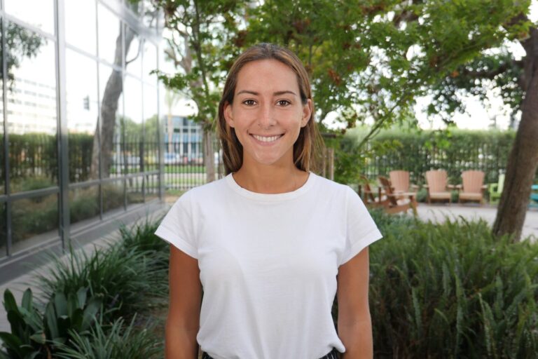 A woman in a white t-shirt stands outside in front of greenery, smiling at the camera, with trees and outdoor seating visible in the background.
