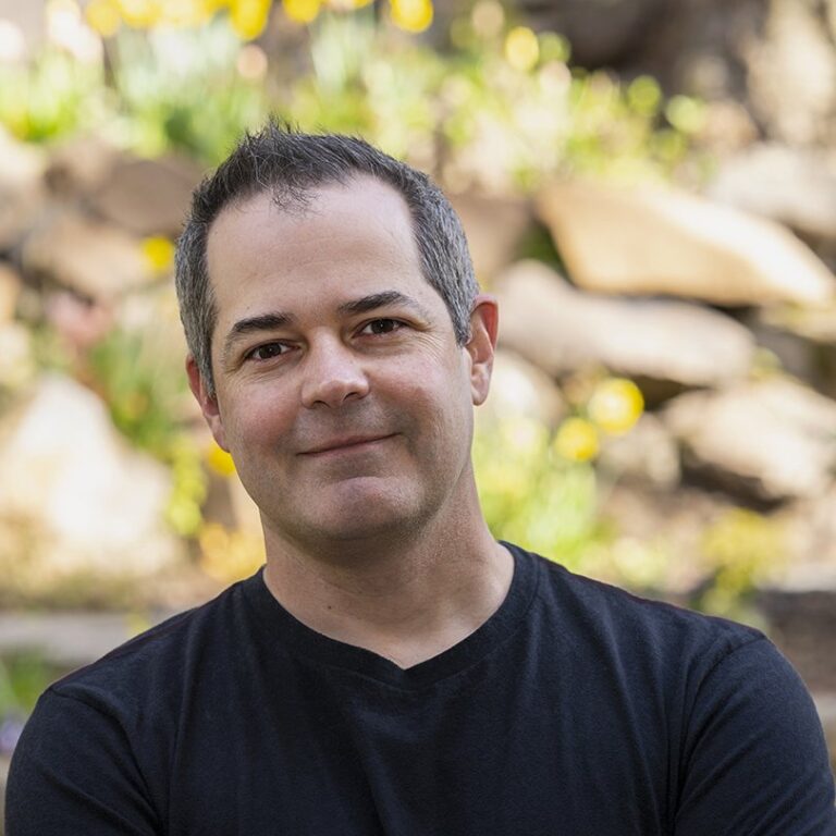 A man with short dark hair and a black t-shirt stands outdoors in front of blurred rocks and greenery, looking at the camera.
