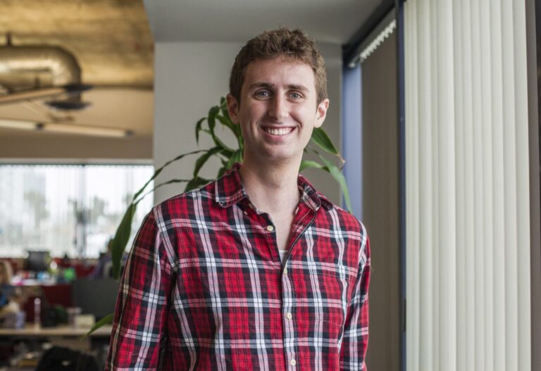 A young man in a red plaid shirt stands indoors near a window with vertical blinds, smiling at the camera. An office setting is visible in the background.