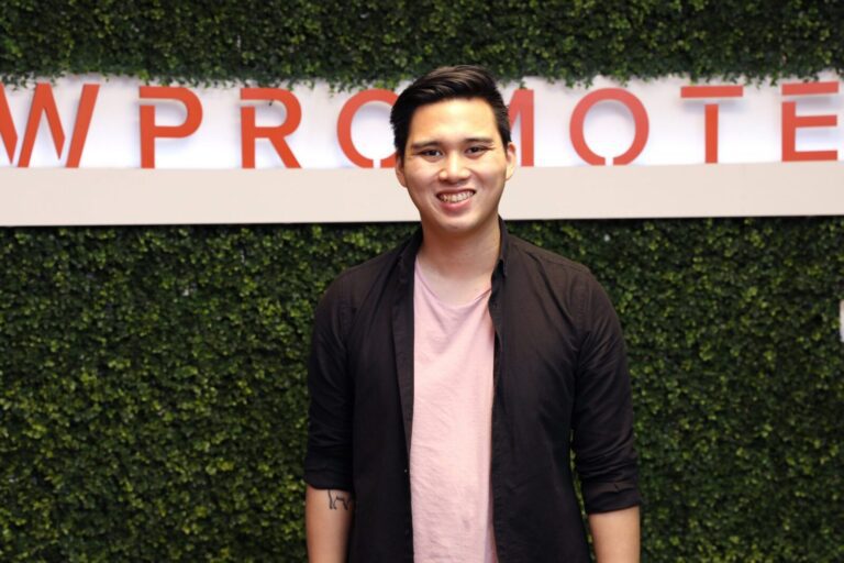 A person stands smiling in front of a green leafy wall with partially visible red text that reads "W PROMOTE.