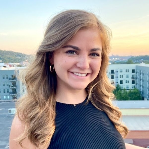 A young woman with long wavy hair, wearing a sleeveless black top and hoop earrings, smiles outdoors with buildings and trees in the background at sunset.