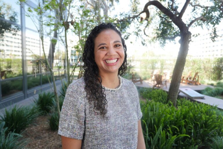 A woman with curly hair, wearing a short-sleeved gray top, stands smiling outdoors near green plants and trees, with buildings and a wooden seating area in the background.