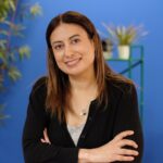 A woman with long brown hair in a black top stands with arms crossed, smiling in front of a blue background with green plants.
