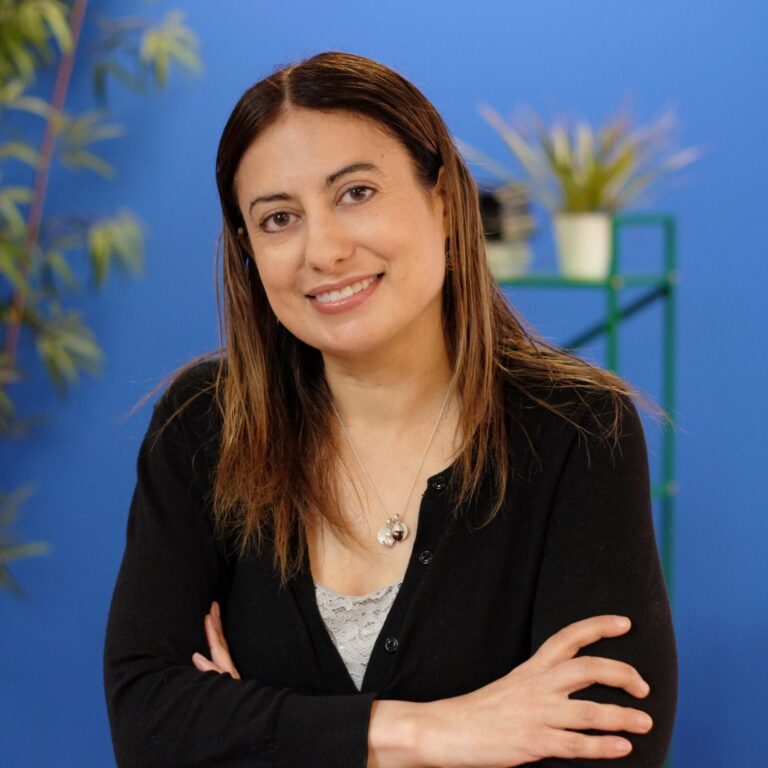 A woman with long brown hair in a black top stands with arms crossed, smiling in front of a blue background with green plants.