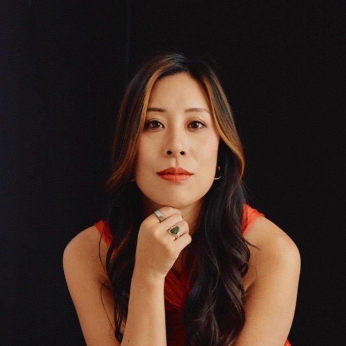A woman with long dark hair and a red dress poses against a black background, resting her chin on her hand and looking directly at the camera.