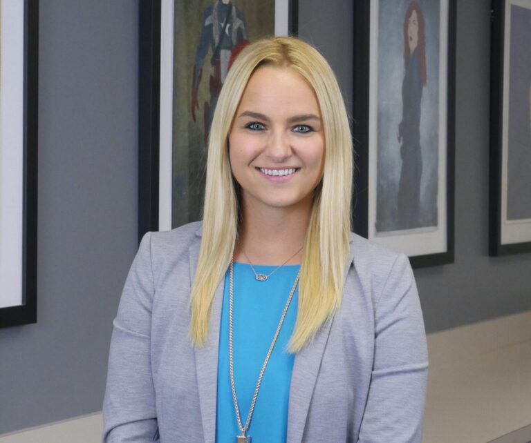 A woman with long blonde hair wearing a light gray blazer and blue top stands indoors in front of framed artwork on a wall, smiling at the camera.