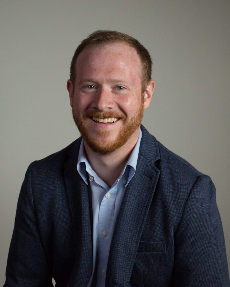 A man with short red hair and a beard, wearing a blue blazer and light blue shirt, smiles while seated against a plain background.