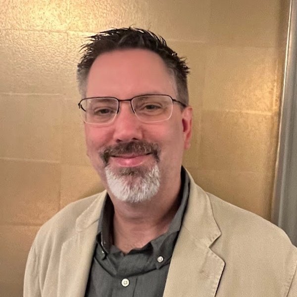 A man with short dark hair, glasses, and a goatee wearing a beige blazer over a dark shirt stands in front of a gold-toned wall, smiling at the camera.