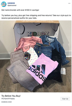 A box containing folded children's clothing, including jeans, a denim jacket, a pink Adidas shirt, a Guess shirt, and a polka-dotted top, displayed in front of a Kidbox box.