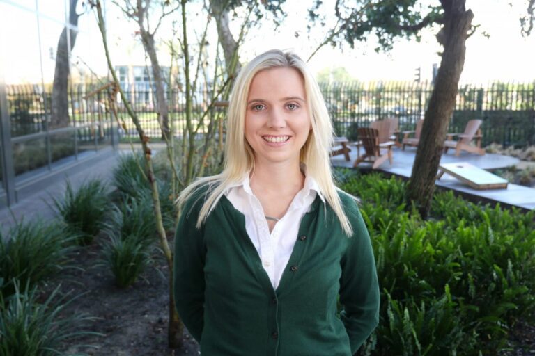 A woman with long blonde hair, wearing a green cardigan over a white shirt, stands and smiles in an outdoor garden area with trees, plants, and wooden chairs in the background.