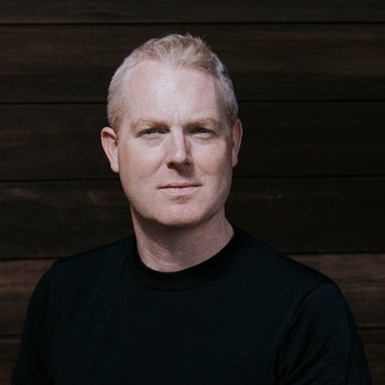 A man with short blond hair wearing a black shirt stands in front of a dark wooden wall, looking directly at the camera.