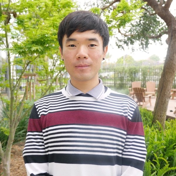 A person with short black hair stands outdoors in front of greenery, wearing a black, white, and red striped sweater.